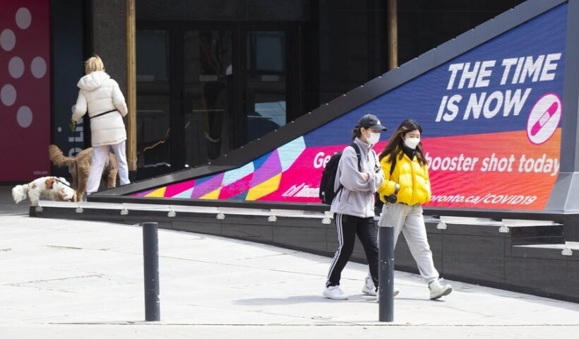 People wearing face masks walk past a screen displaying the COVID-19 vaccination information in Toronto
