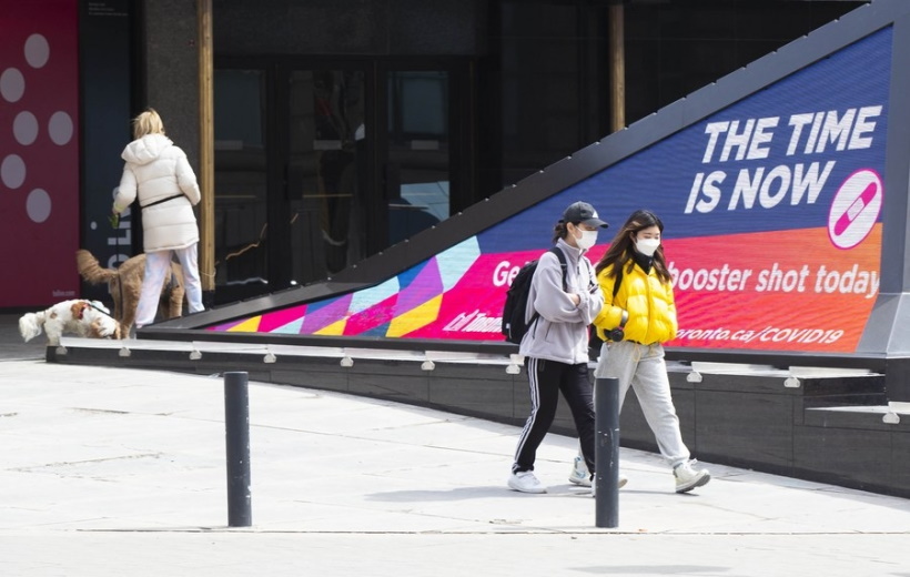 People wearing face masks walk past a screen displaying the COVID-19 vaccination information in Toronto